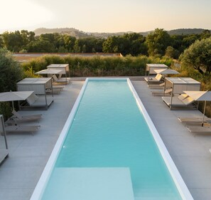 A rectangular swimming pool with a white border, surrounded by lounge chairs and umbrellas, set against a backdrop of trees and hills. - Gandum Village - Conscious Boutique Hotel (Montemor-o-Novo)