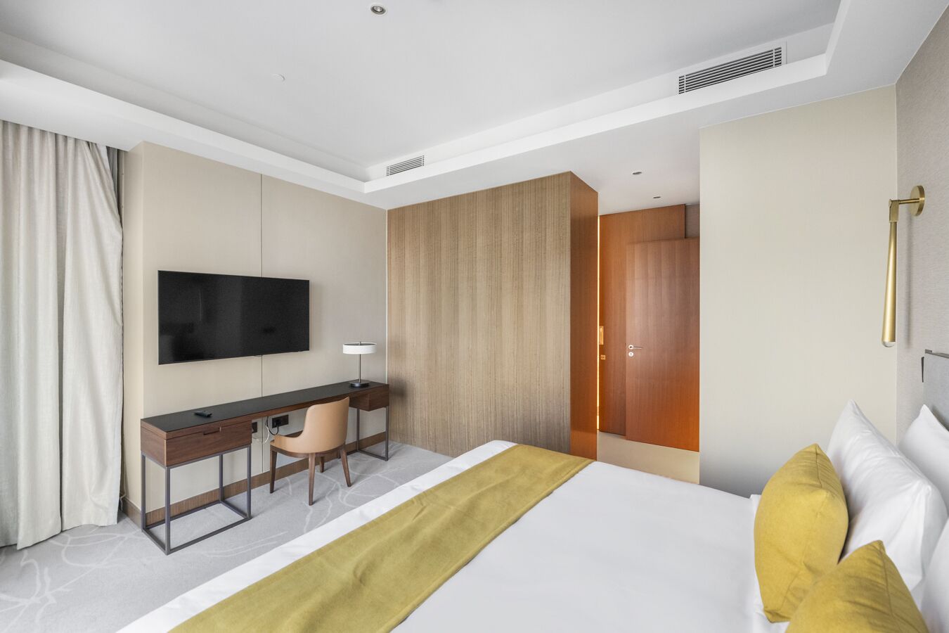 A modern hotel room with a bed, a desk, a chair, a television, and a wardrobe.