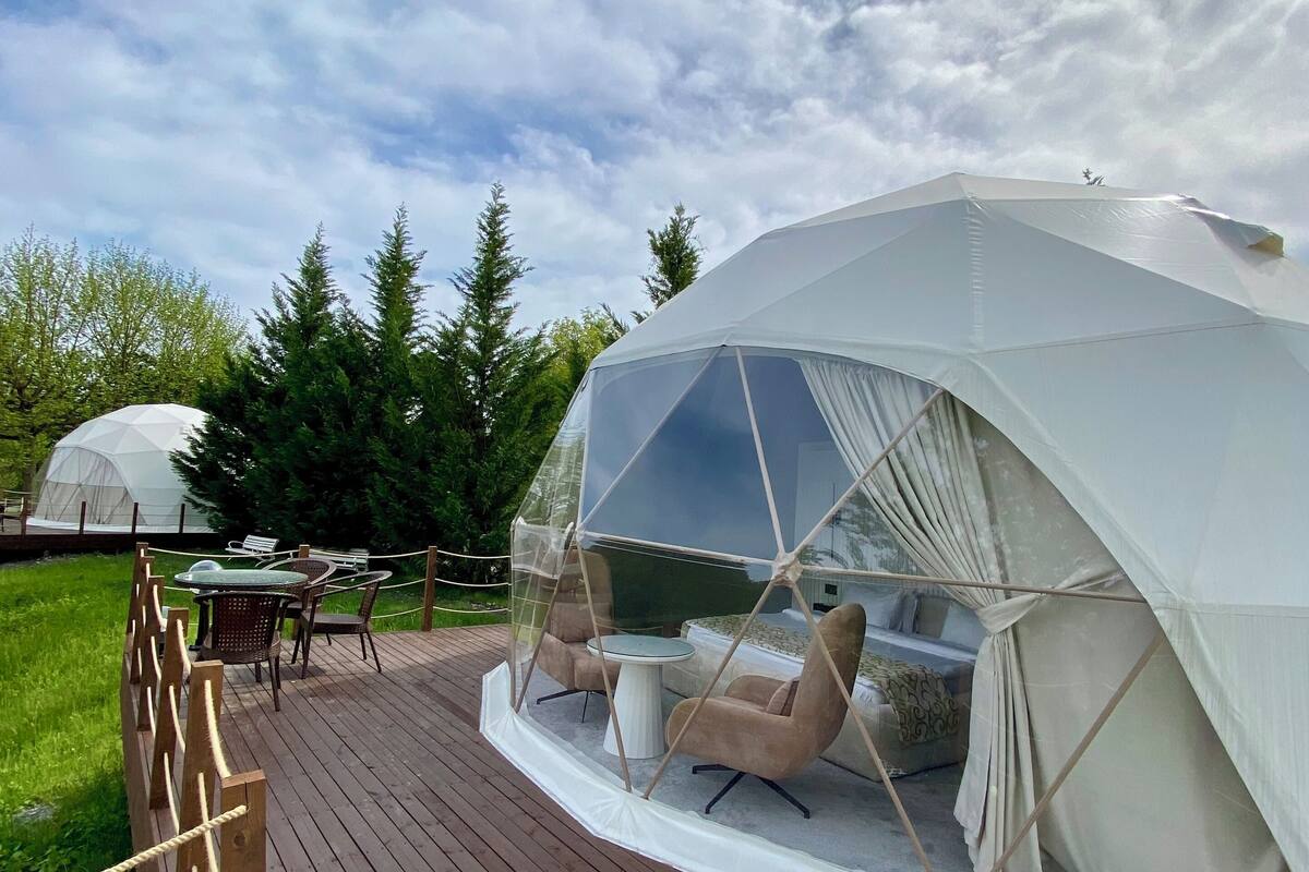 A dome-shaped tent with a transparent ceiling, furnished with a sofa and a table, set on a wooden deck with a rope barrier.