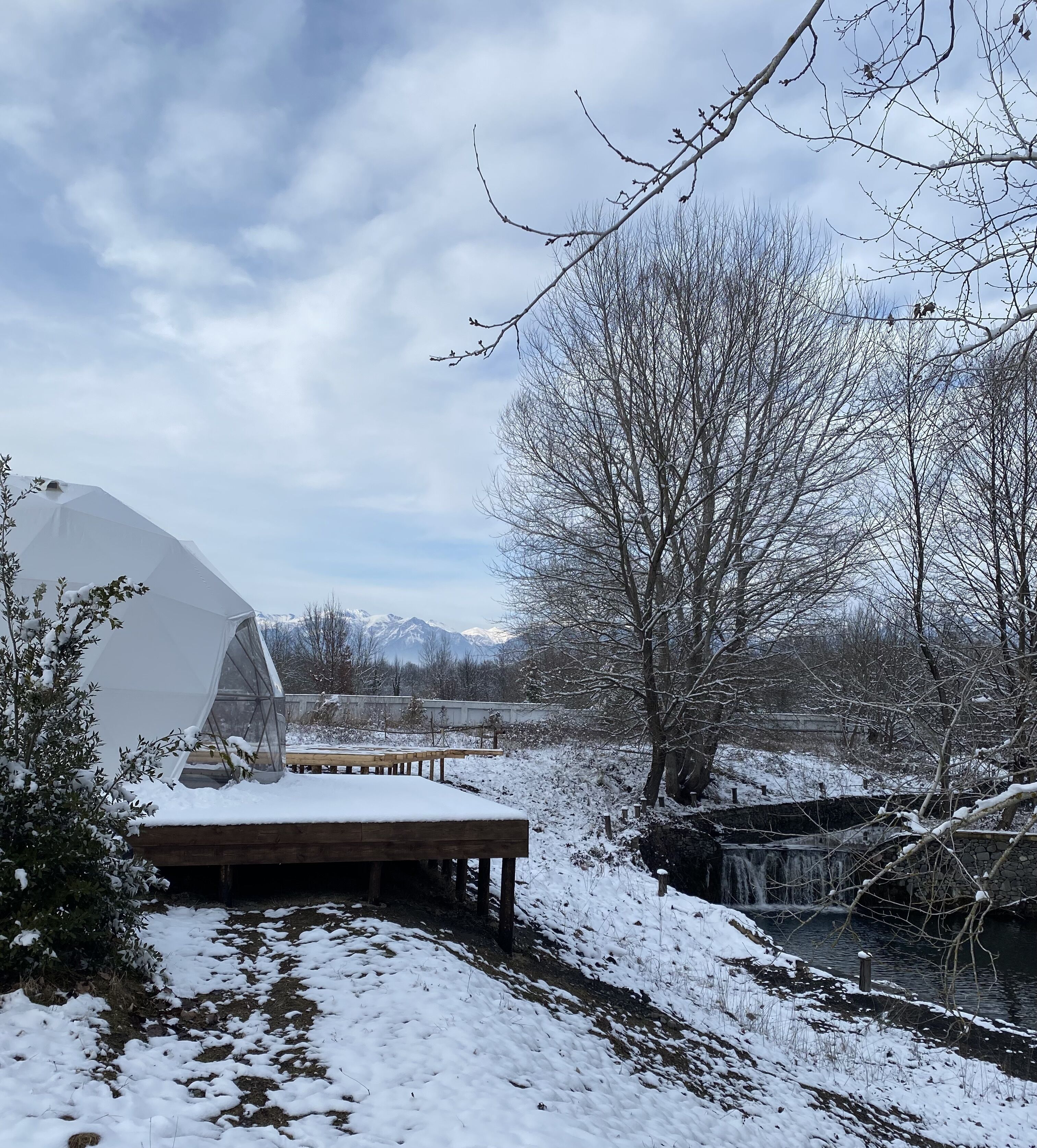 A snow-covered outdoor area with a wooden deck, a greenhouse, and a view of mountains in the background.