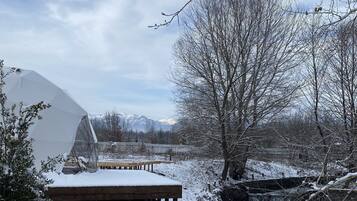 A snow-covered outdoor area with a wooden deck, a greenhouse, and a view of mountains in the background.