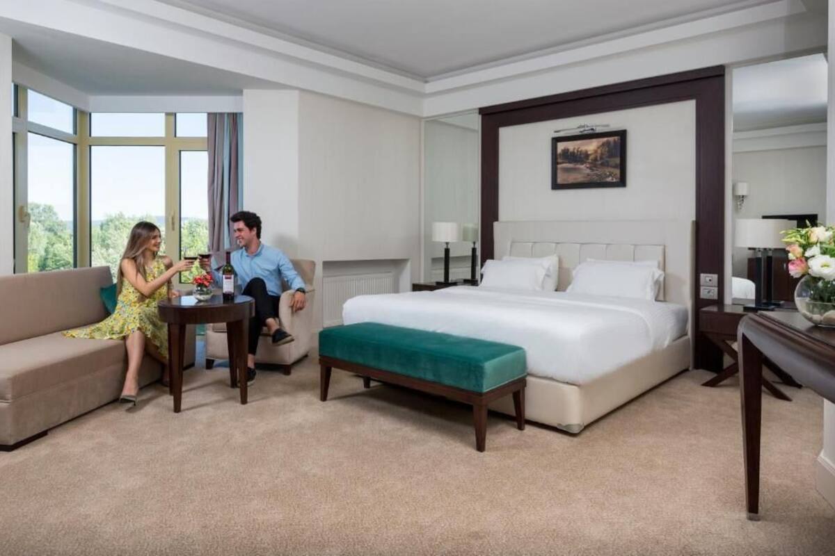 A hotel room with a large bed, a sofa, a small table, and a view of the outdoors.