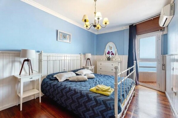A bedroom with a bed, bedside tables, a dresser, a mirror, and a view of the sea.