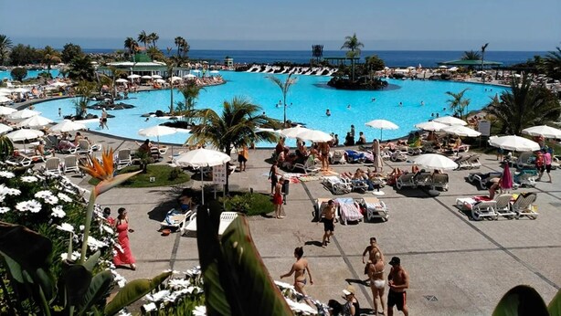 Ein großer Pool im Freien, umgeben von Liegestühlen und Sonnenschirmen, mit Palmen und freiem Blick auf das Meer.
