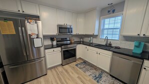 A modern kitchen with white cabinets, stainless steel appliances, a microwave, and a window with blinds. - Pelican's Loft: Steps from the beach access  (Carolina Beach)