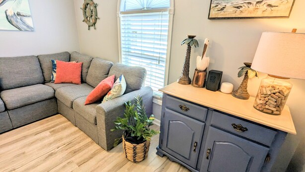 A living room with a gray sectional sofa, a wooden floor, a blue cabinet, and a window with blinds. - Pelican's Loft: Steps from the beach access  (Carolina Beach)