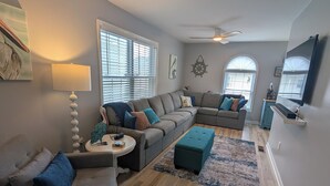 A modern living room with a sectional sofa, a round coffee table, and a teal ottoman. There are two ceiling fans, one with a wooden blade and one with a white blade. The room has a large window with blinds, a wall-mounted TV, and a decorative wall piece. - Pelican's Loft: Steps from the beach access  (Carolina Beach)
