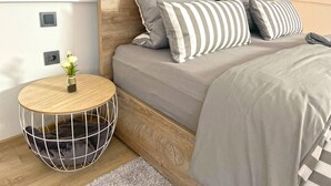 Une chambre moderne avec une tête de lit en bois, une literie grise et une petite table d’appoint ronde sur laquelle est posé un vase de fleurs.