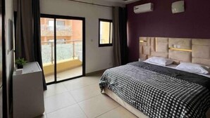 A modern hotel room with a large bed, a small desk, and a sliding glass door to the balcony. - La Liga, 2 Bedrooms (Abidjan)