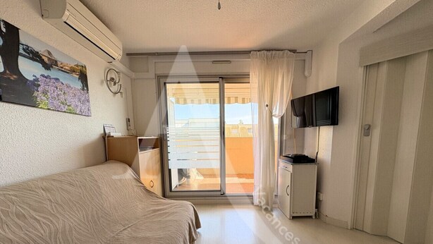 A bedroom with a bed, a TV, a desk, and a sliding glass door to a balcony. - T2, 2ème sans ascenseur, garage, crique de Lanau (Sète)