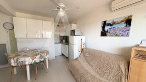 A compact room with a dining table, a sofa, a ceiling fan, a kitchen area, and a wall-mounted air conditioner. - T2, 2ème sans ascenseur, garage, crique de Lanau (Sète)