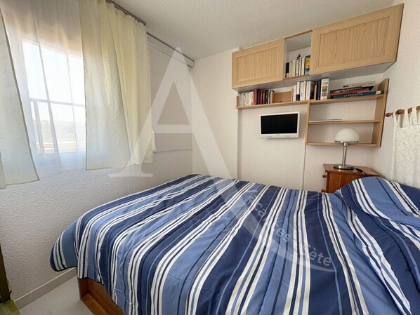 A bedroom with a bed, a TV, a bookshelf, and a window with curtains. - T2, 2ème sans ascenseur, garage, crique de Lanau (Sète)