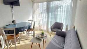 A living room with a sofa, a dining table with chairs, a small table with a plant, and a TV mounted on the wall. - T3 avec balcon coeur quartier Corniche (Sète)