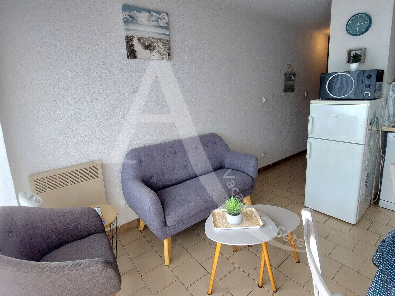 A compact living space with a grey sofa, a small round table with a plant, a white chair, and a white refrigerator.