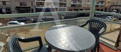 Un balcon avec une table ronde et deux chaises, donnant sur une rue bordée de voitures garées et sur laquelle on aperçoit des bâtiments au loin.