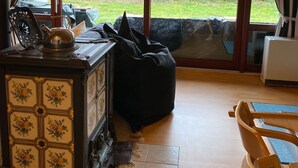 A cozy room with a wooden ceiling, a large window with curtains, a wooden cabinet with floral patterns, a black bean bag, and a wooden chair.