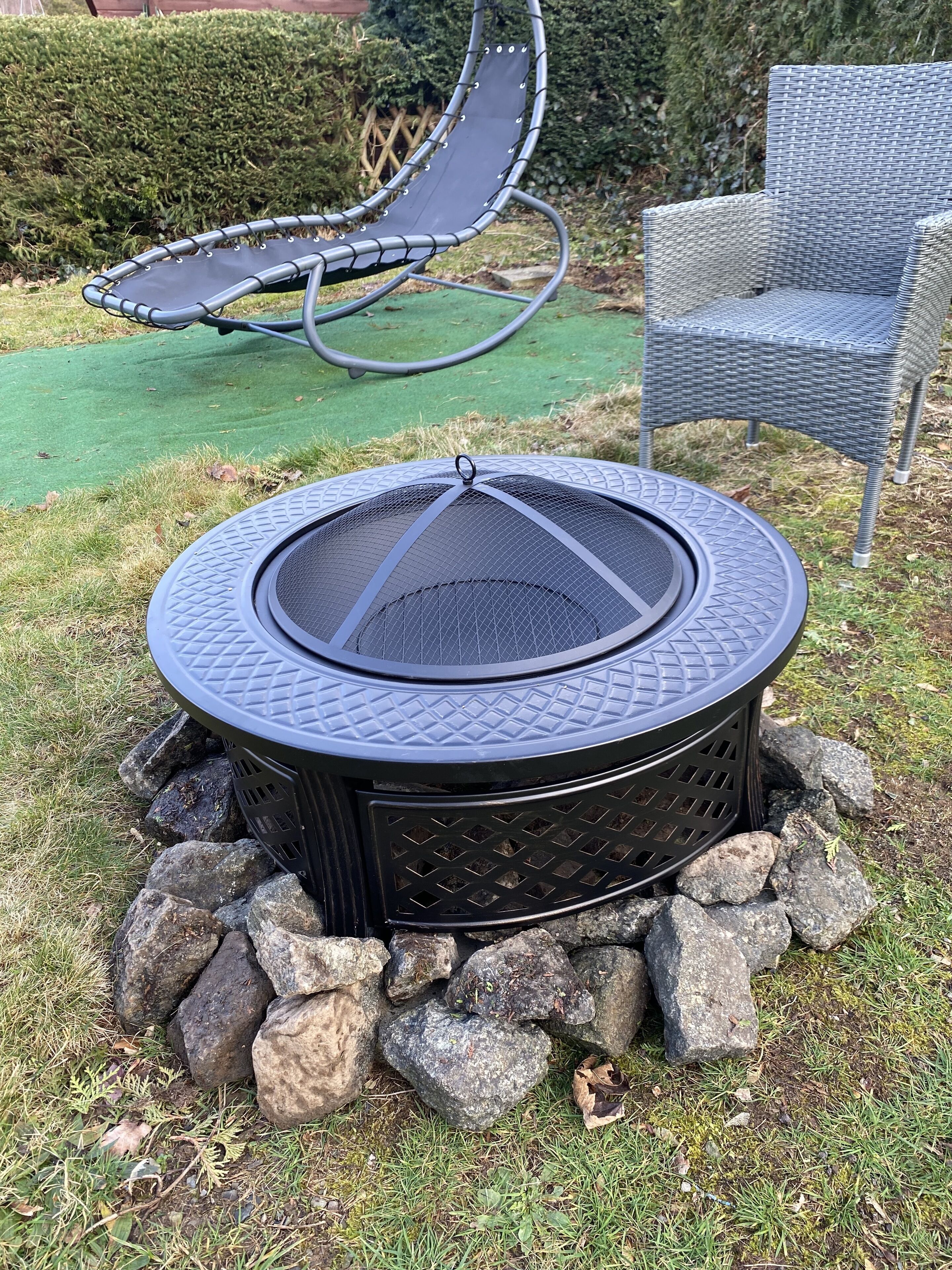 Een ronde vuurkuil buiten met een metalen grill, omringd door rotsen, in een grasrijke omgeving met een stoel en een hangmat op de achtergrond.
