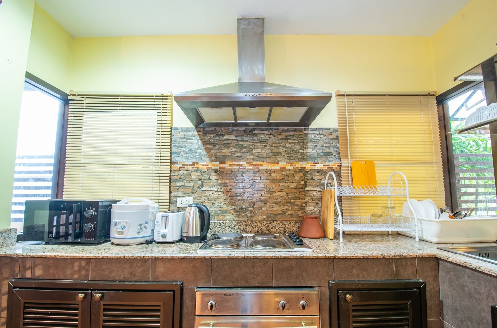 Una cocina con protector mural de piedra, campana extractora de acero inoxidable y horno empotrado. Hay varios electrodomésticos en la encimera, incluyendo un microondas, tostadora y cafetera.