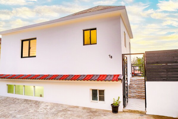 Un bâtiment moderne à deux étages, doté d’une façade blanche et portant le numéro 18, se distingue par son toit recouvert de tuiles rouges, un petit pot de fleurs et une entrée fermée par un portail.