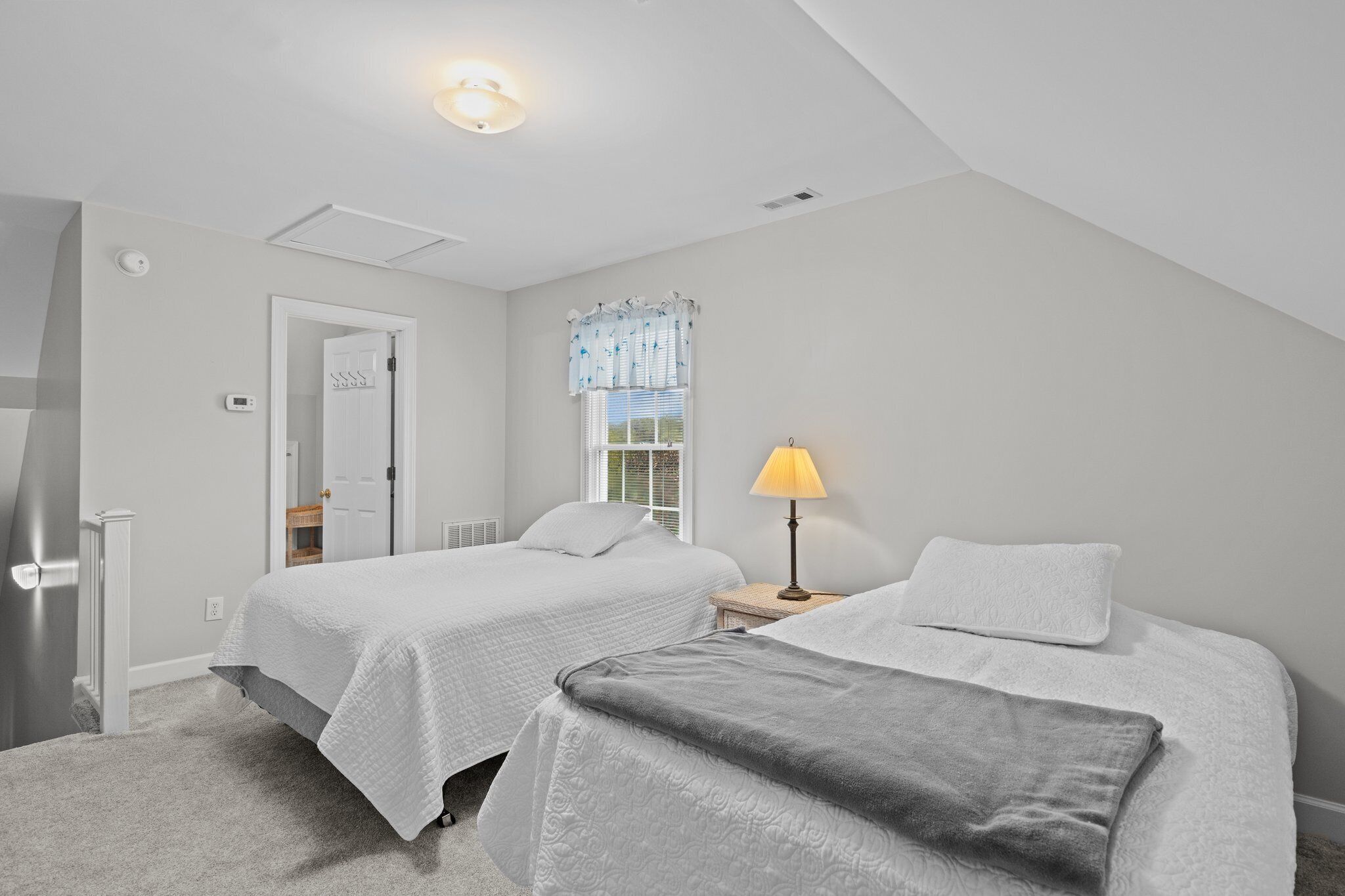 A bedroom with two beds, a nightstand with a lamp, a window with blinds, and a door leading to another room.