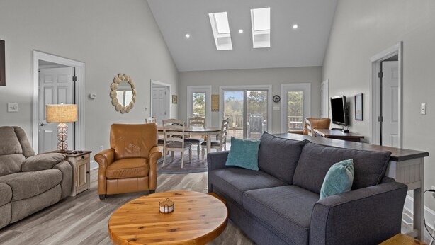 A spacious living room with a gray sofa, a brown leather chair, and a wooden coffee table. There is a dining area with a table and chairs, and a kitchen area with a counter and a television. - Red Buffalo| Steps to beach and town. (Emerald Isle)