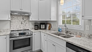 A kitchen with white cabinets, a stainless steel stove, a coffee maker, and a window with blinds. - Red Buffalo| Steps to beach and town. (Emerald Isle)