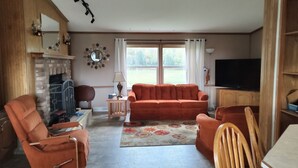 Olohuone, jossa on takka, oranssi sohva, puutuolit, televisio ja suuri ikkuna, jossa on verhot.