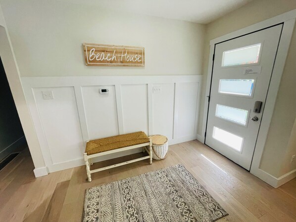 Ein heller, minimalistischer Flur mit einer Bank, einem gemusterten Teppich und einem Schild mit der Aufschrift 'Beach House'.