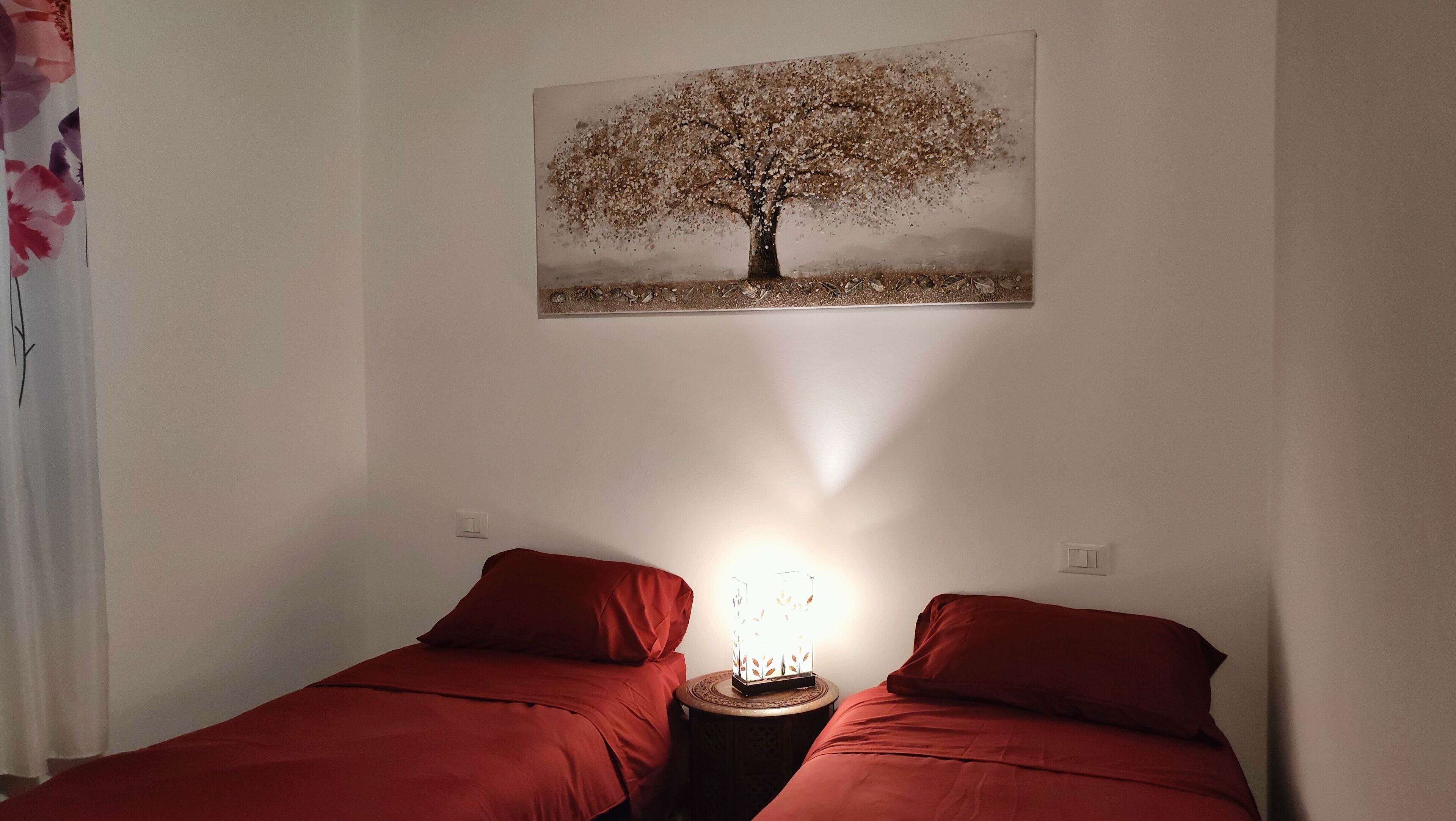 Una camera con due letti singoli, un comodino con una lampada e un quadro di un albero appeso alla parete.