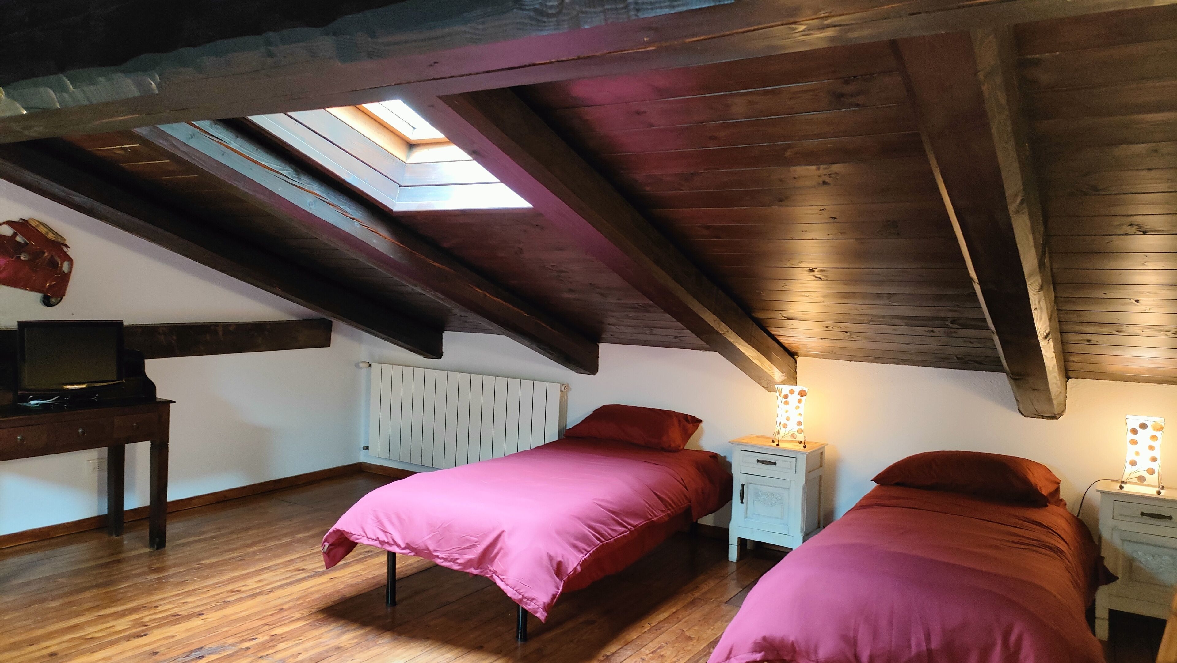 Une chambre avec deux lits, un plafond en bois, un puits de lumière, une table de chevet avec une lampe et un téléviseur posé sur une commode.