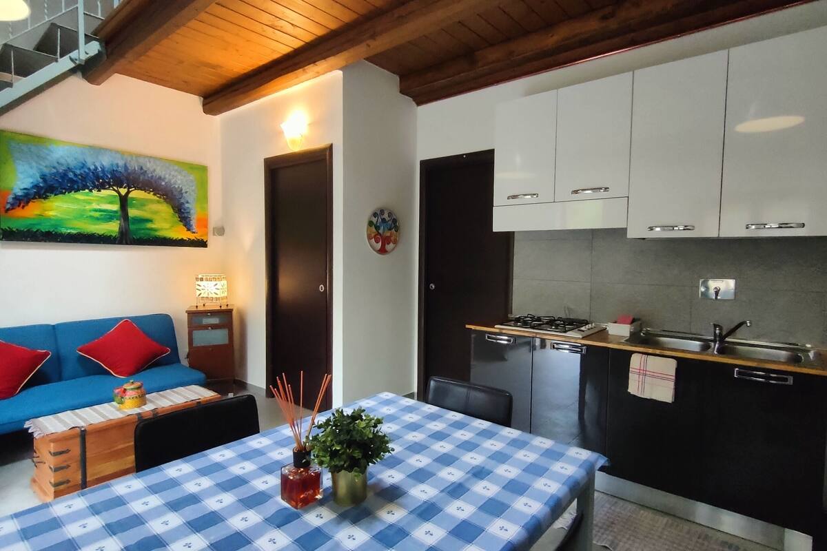 A compact kitchen with white cabinets, a blue and white checkered tablecloth, and a painting of a tree on the wall.