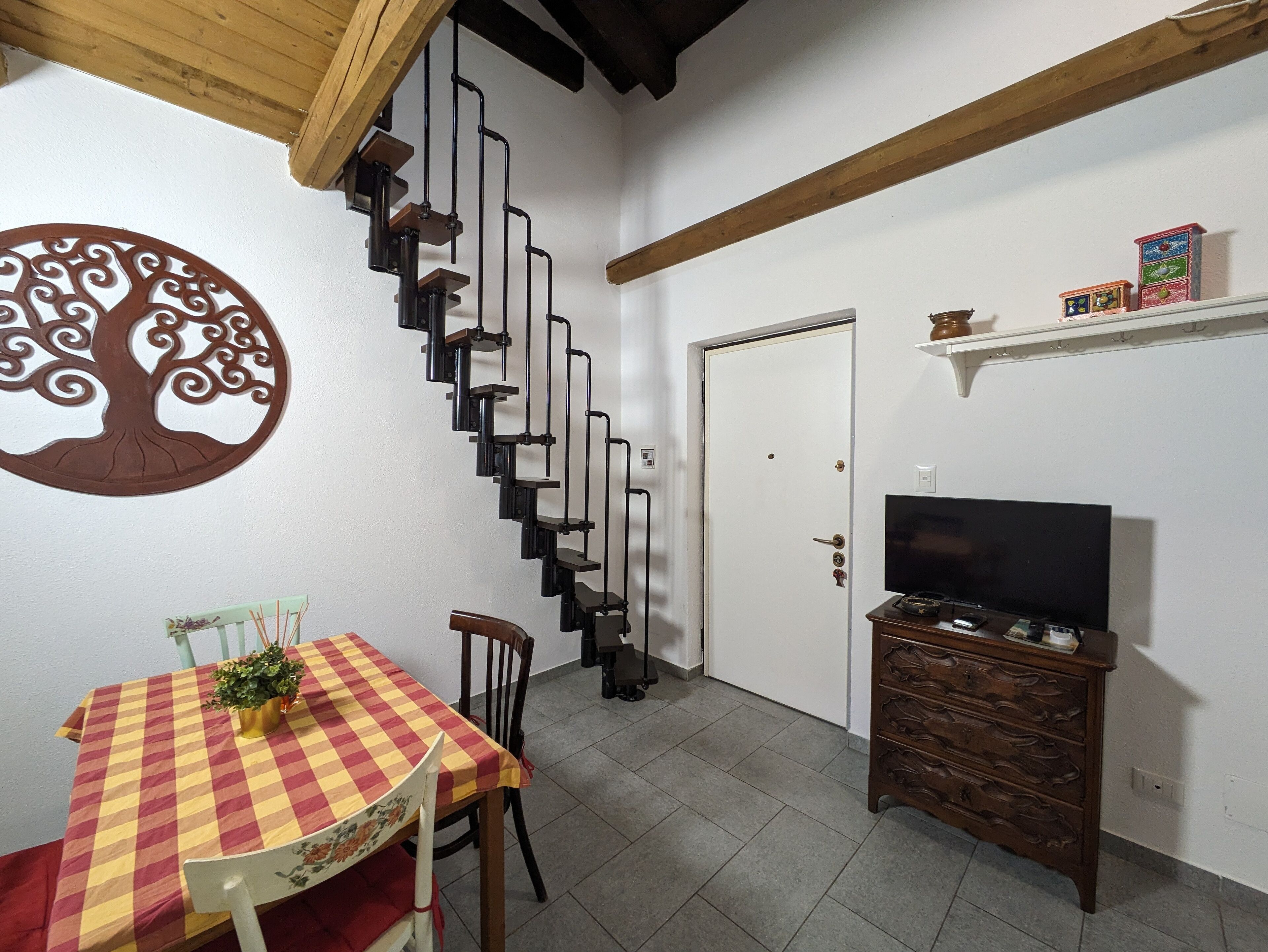A dining area with a checkered tablecloth, a chair, a small potted plant, and a staircase with a tree wall art piece.