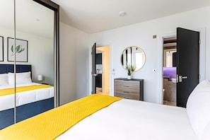 Ein Hotelzimmer mit einem Bett, einem Nachttisch, einem Spiegel, einer Kommode und einer offenen Tür, die in einen weiteren Raum führt.