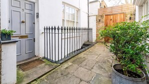 Une ruelle étroite, pavée de pierres, avec une porte blanche, une grille en fer forgé noire et, au fond, une porte en bois.