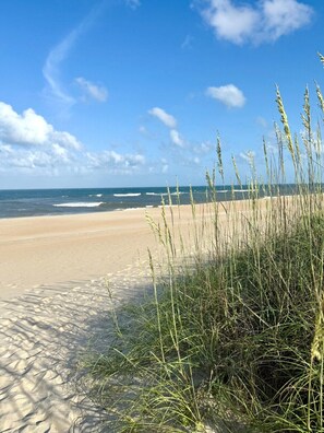 Praia com areia, ondas e grama alta em primeiro plano.