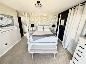 Uma cama de dossel com cortina, uma mesa de cabeceira com abajur, uma cômoda com gavetas e um quadro na parede.