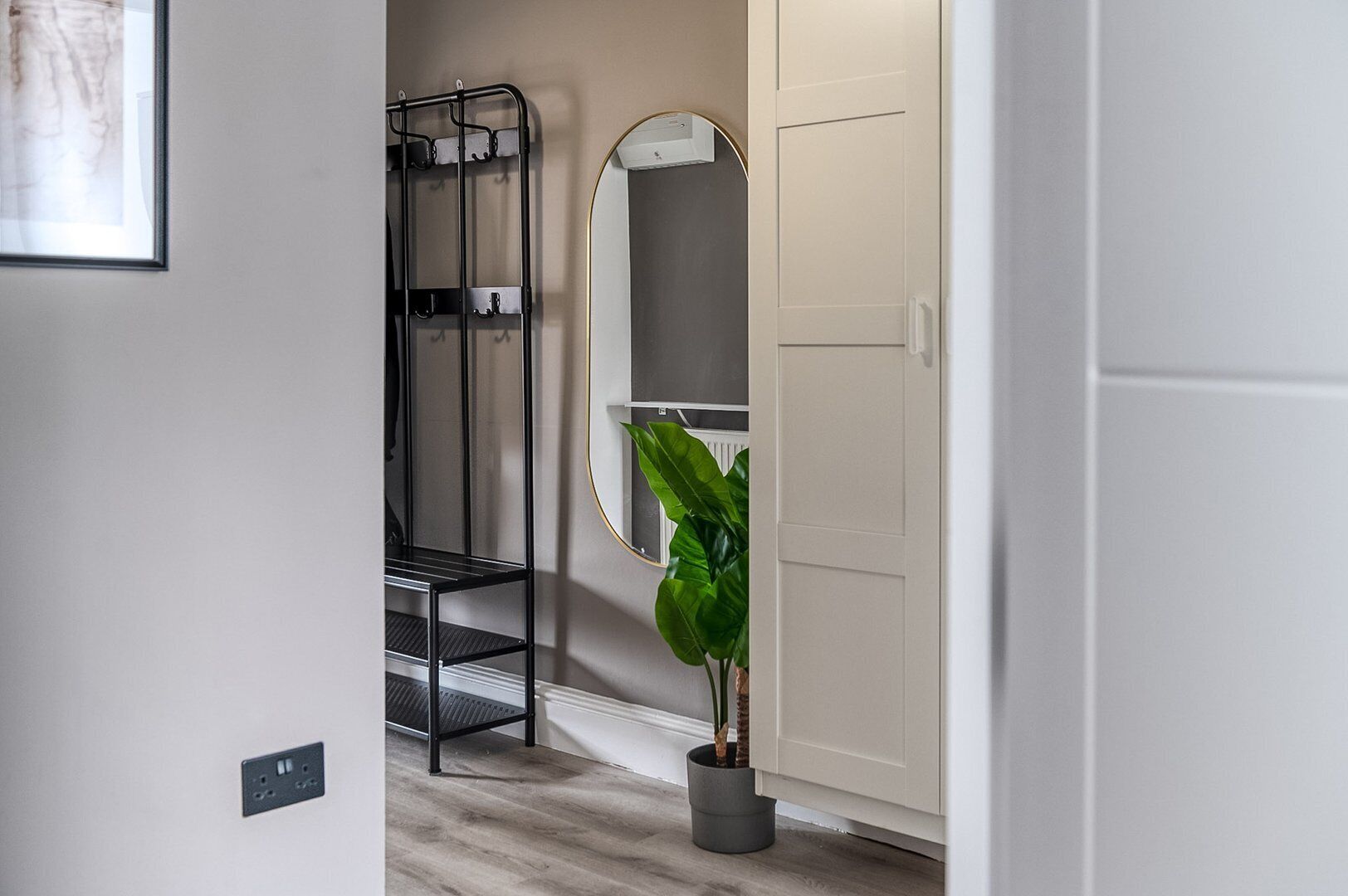 Un pasillo con un estante de metal negro, un espejo, una planta en maceta y una puerta color beige.