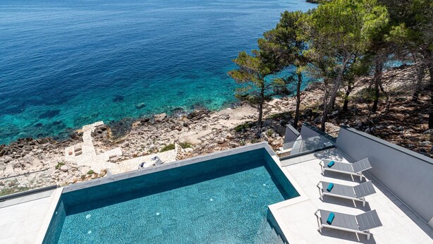 Une piscine à l’eau d’un bleu cristallin, entourée de chaises longues blanches et bordée d’un paysage rocheux parsemé d’arbres.