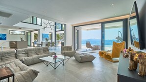 Un vaste espace de vie agrémenté d’un canapé moderne, d’une table basse en verre et offrant une vue sur la mer grâce à de grandes fenêtres.