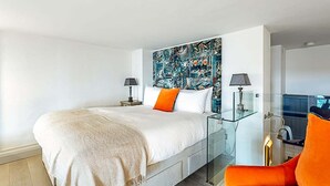 A modern bedroom with a large bed, glass bedside tables, and a vibrant abstract wall art.