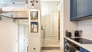 A compact kitchen with a built-in oven, a coffee maker, and a toaster. There are framed pictures on the wall and a staircase leading to a bedroom.