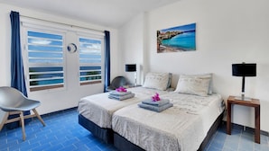 Une chambre d’hôtel comprenant un lit, deux tables de chevet, une chaise, une fenêtre avec des rideaux et un tableau représentant une scène de plage.