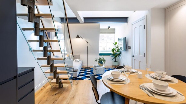 A modern kitchen with a wooden staircase, a dining area with a round table set for two, and a view of the city through large windows. - Coleherne Road Residence IX (London)