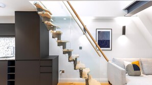 A modern interior with a unique wooden staircase, a grey sofa, and a kitchen area. - Coleherne Road Residence IX (London)