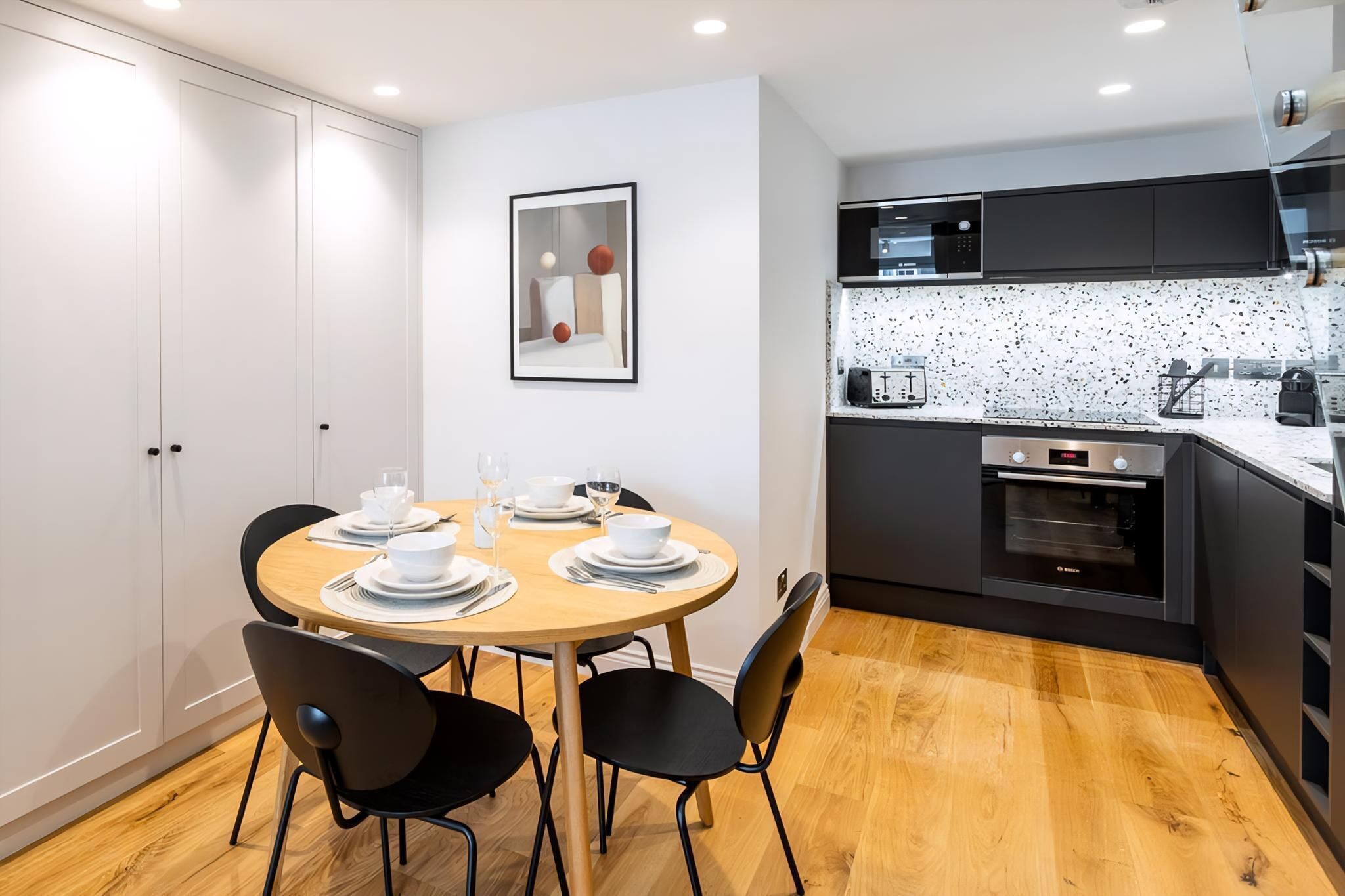A modern kitchen with a microwave, oven, and coffee maker. There is a dining table with chairs and a framed picture on the wall.
