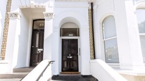 A building entrance with a black door, a number '23' above the door, and a set of stairs leading up to it. - Coleherne Road Residence IX (London)