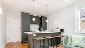 A modern kitchen with a central island, bar stools, pendant lighting, and a green sofa in a living area.