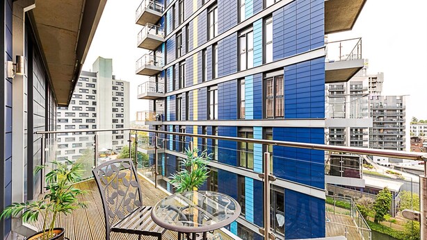 Un balcon doté d’une table et de chaises en verre, de plantes en pot et offrant une vue sur un immeuble moderne à la façade bleue et blanche.