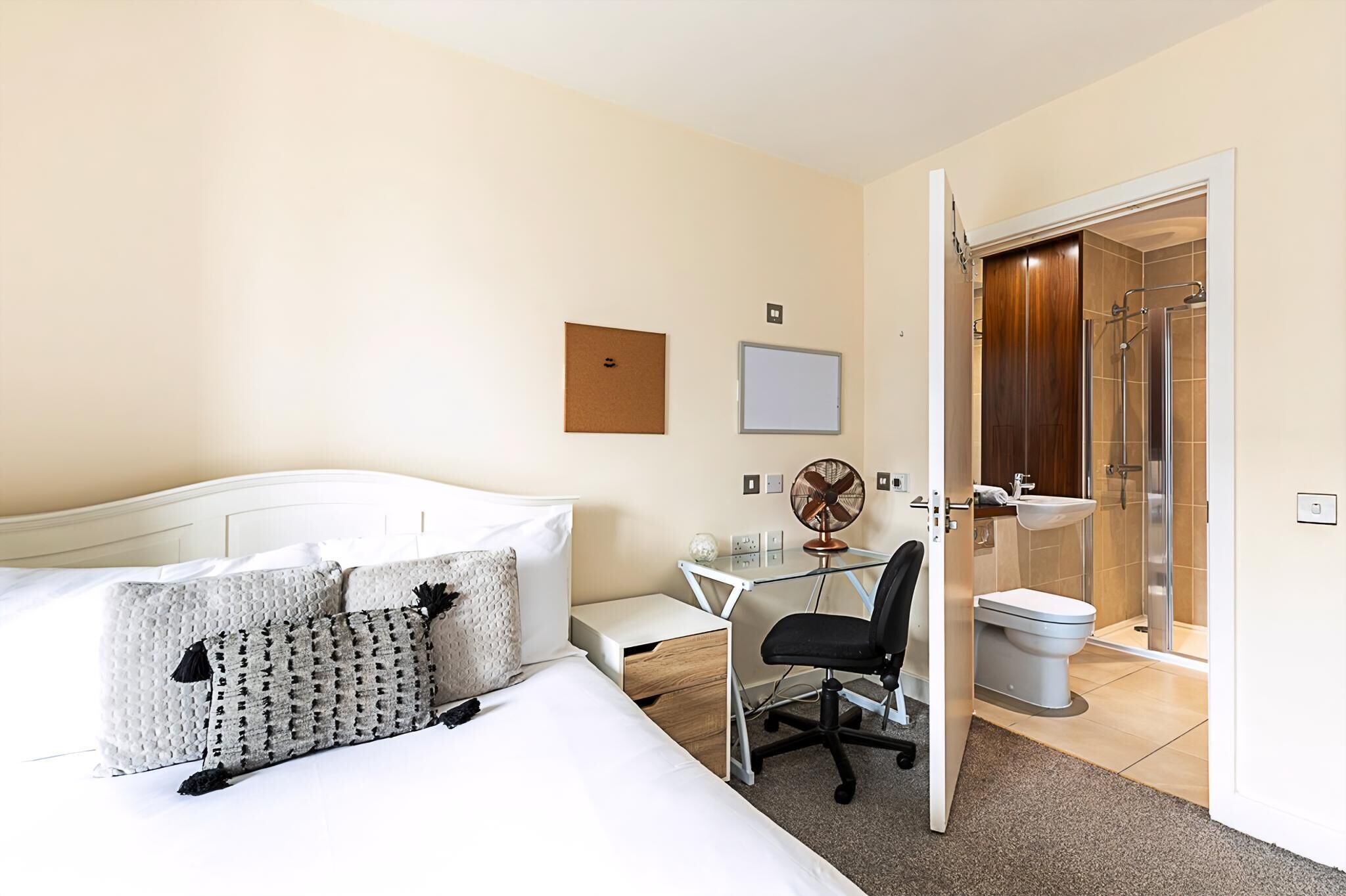 Quarto de hotel com cama, escrivaninha, cadeira, ventilador, pia e chuveiro.