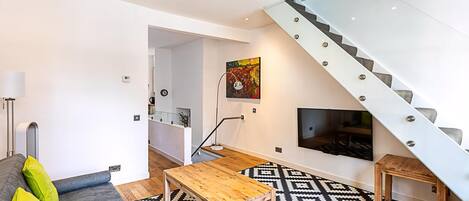A modern living room with a sofa, a coffee table, a patterned rug, a staircase, and a TV mounted on the wall.
